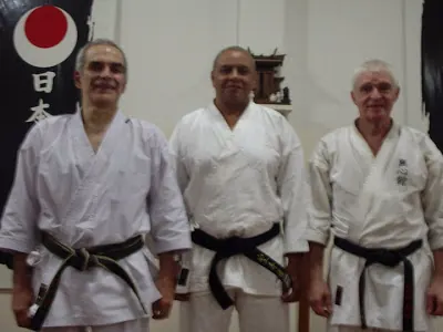 Sensei Carlos Rocha com Ricardo D’Elia e Carlos Garitta, de quimono, diante de um banner do dojo