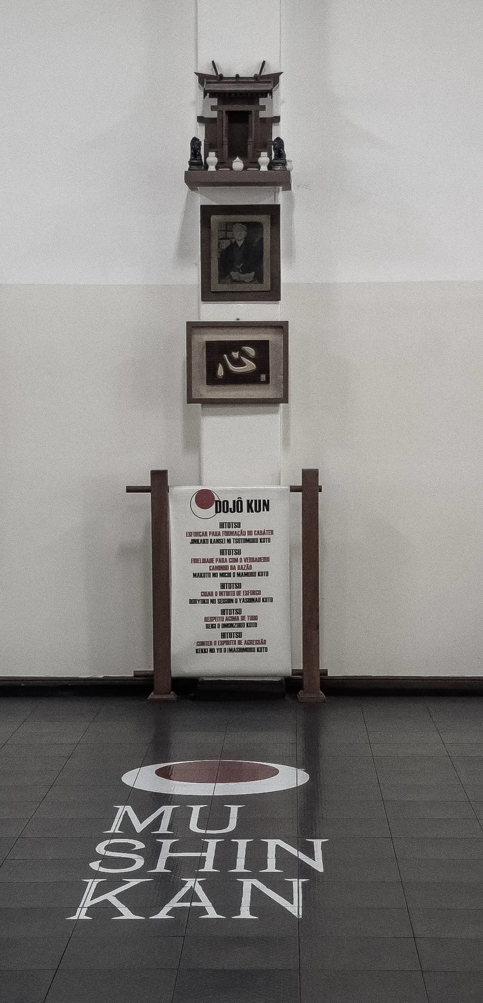 Shomen do dojo: o altar, o retrato de Gichin Funakoshi e o quadro com o Dojo Kun, acima do nome Mushinkan pintado no piso